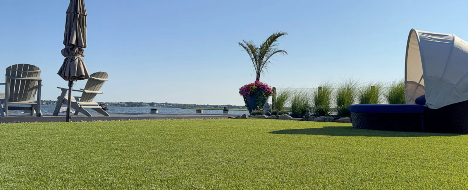 Waterfront lawn with Adirondack chairs, closed umbrella, palm plant, flowers, and shaded outdoor lounge under clear sky.