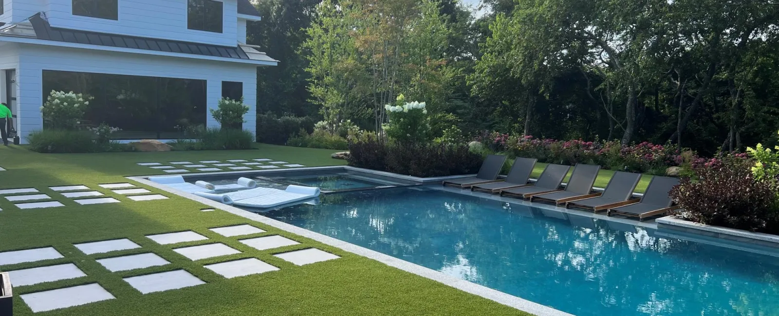 Modern backyard with a swimming pool, lounge chairs, green lawn, and a two-story white house under a partly cloudy sky