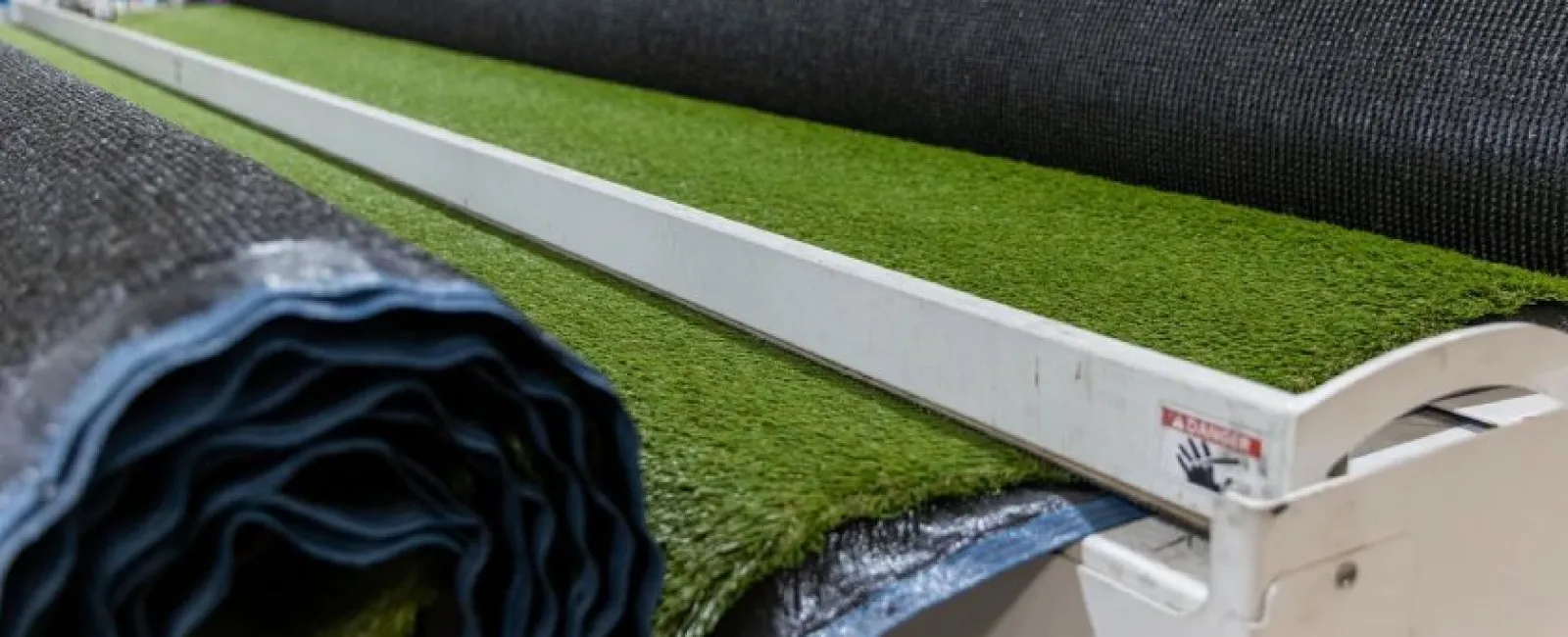 Rolls of artificial green turf on a cutting machine in a warehouse with industrial shelving in the background