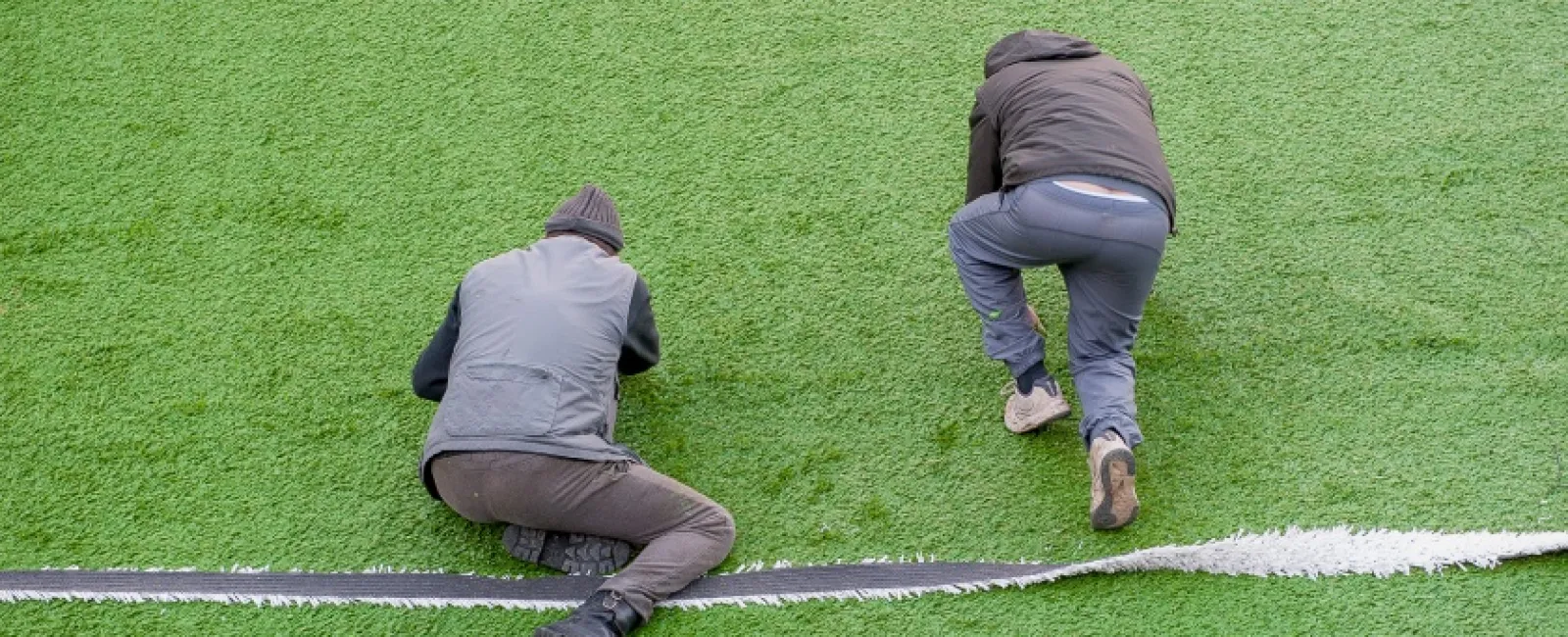 Two workers install artificial turf by aligning and securing the turf edges on a sports field.