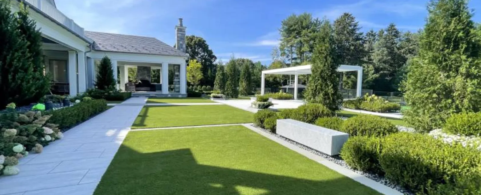Modern backyard with manicured lawn, white stone pathways, pergola, and lush green trees under a clear blue sky