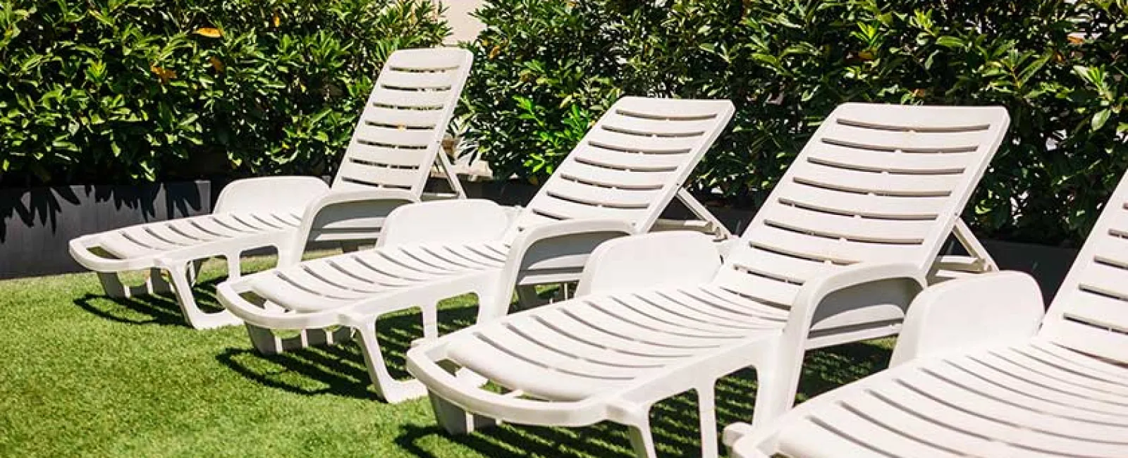 Four white plastic lounge chairs on artificial grass with green bushes and a tiled wall background.