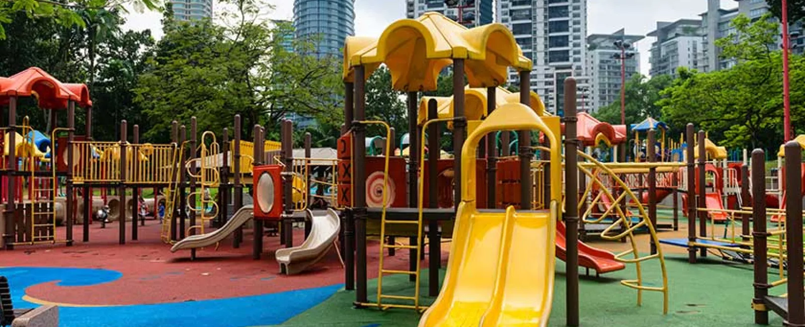 Colorful urban playground with slides, climbing structures, and safety flooring under tall city buildings.