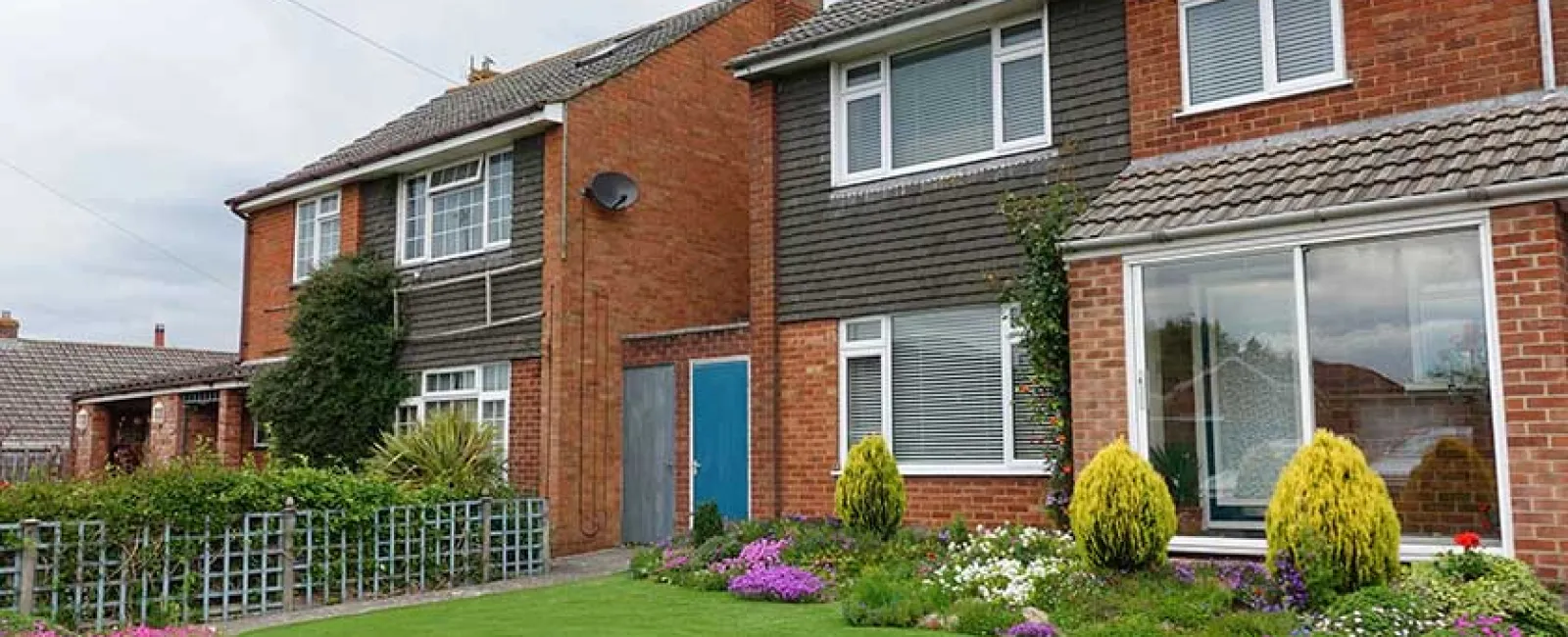 Two attached red brick suburban houses with well-maintained lawns, flowerbeds, and clear windows on a cloudy day