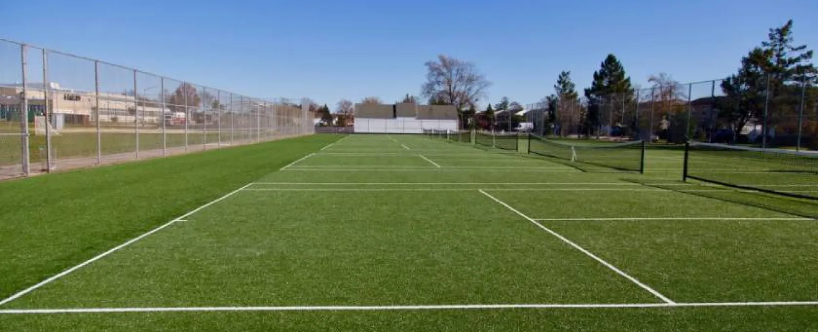 Empty outdoor tennis courts with artificial turf under clear blue sky surrounded by fencing and trees.