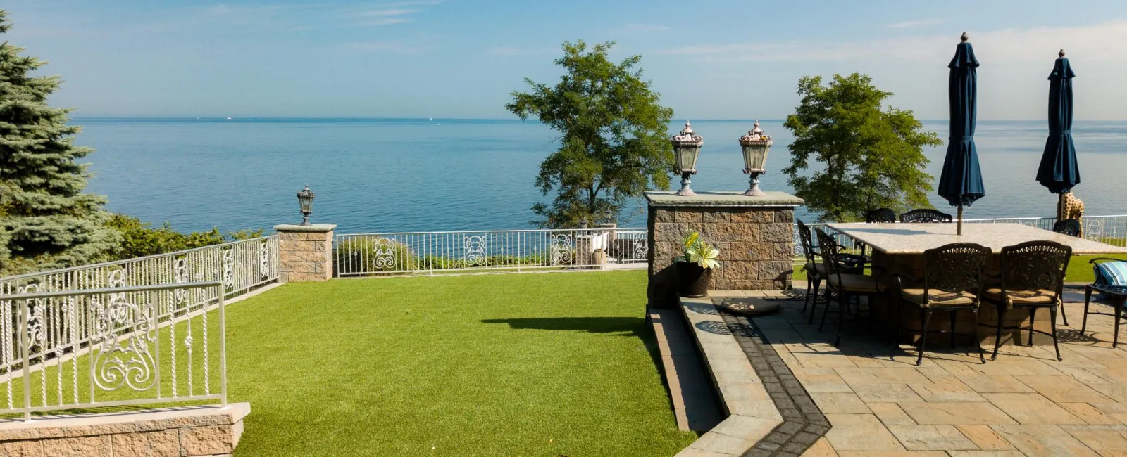 Outdoor patio with dining table, umbrellas, and green lawn overlooking calm blue lake under clear sky