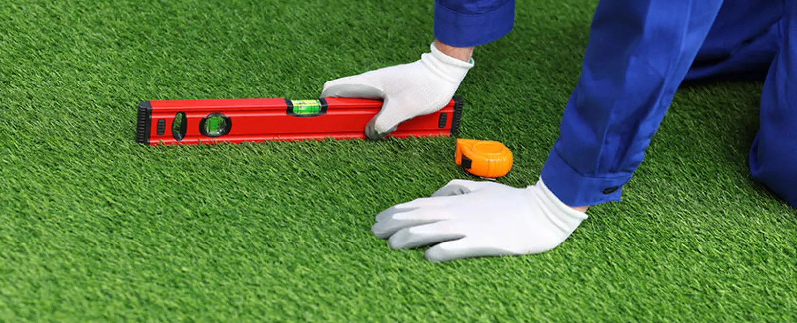 Worker wearing gloves using a red level tool on green artificial grass with measuring tape nearby