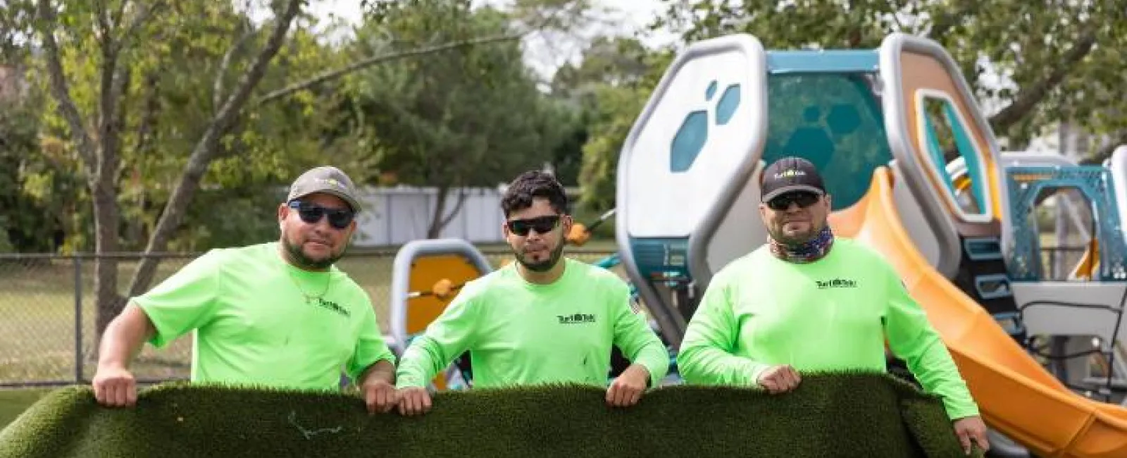 Three workers in neon green shirts holding a rolled artificial grass turf at a playground with slides in background.