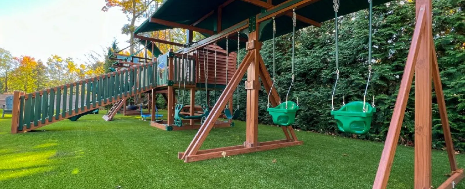 Children's wooden playground set with swings, slides, and climbing structures on green grass under autumn trees.