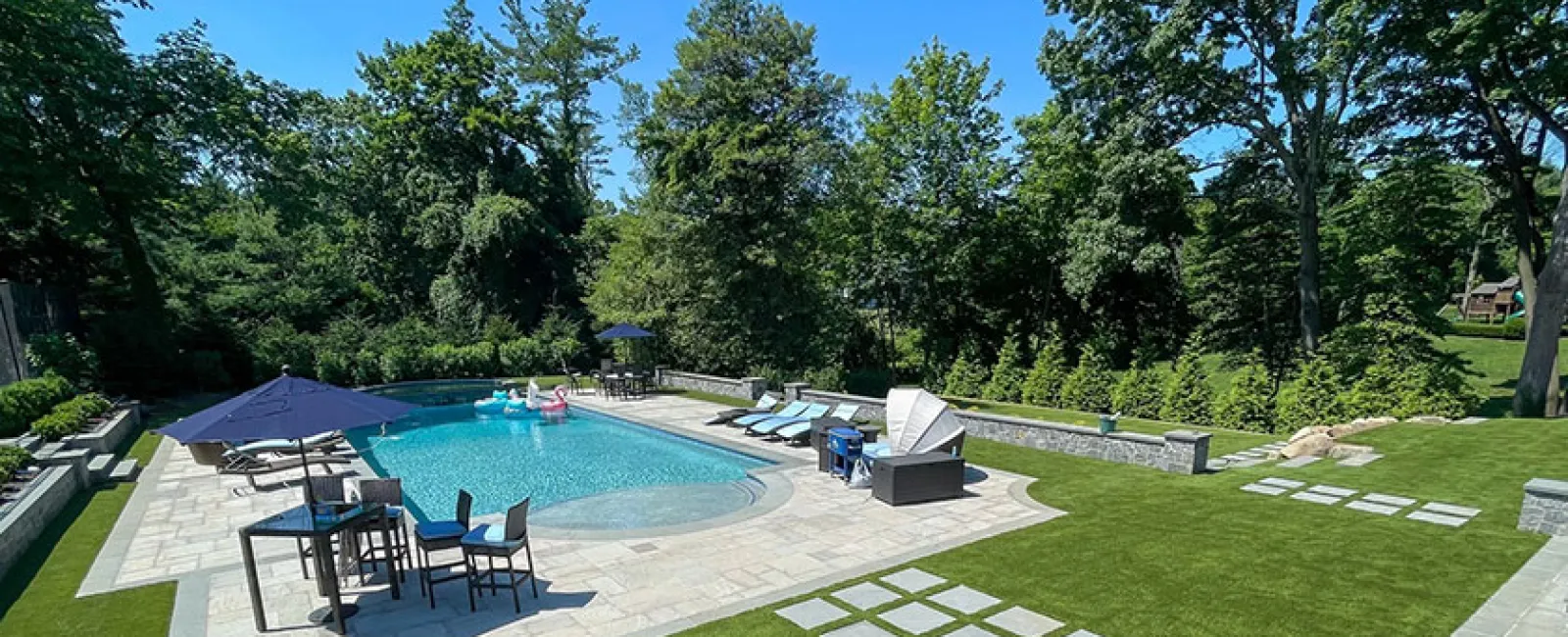 Luxury backyard pool with lounge chairs, umbrellas, seating area, and lush green lawn surrounded by trees.