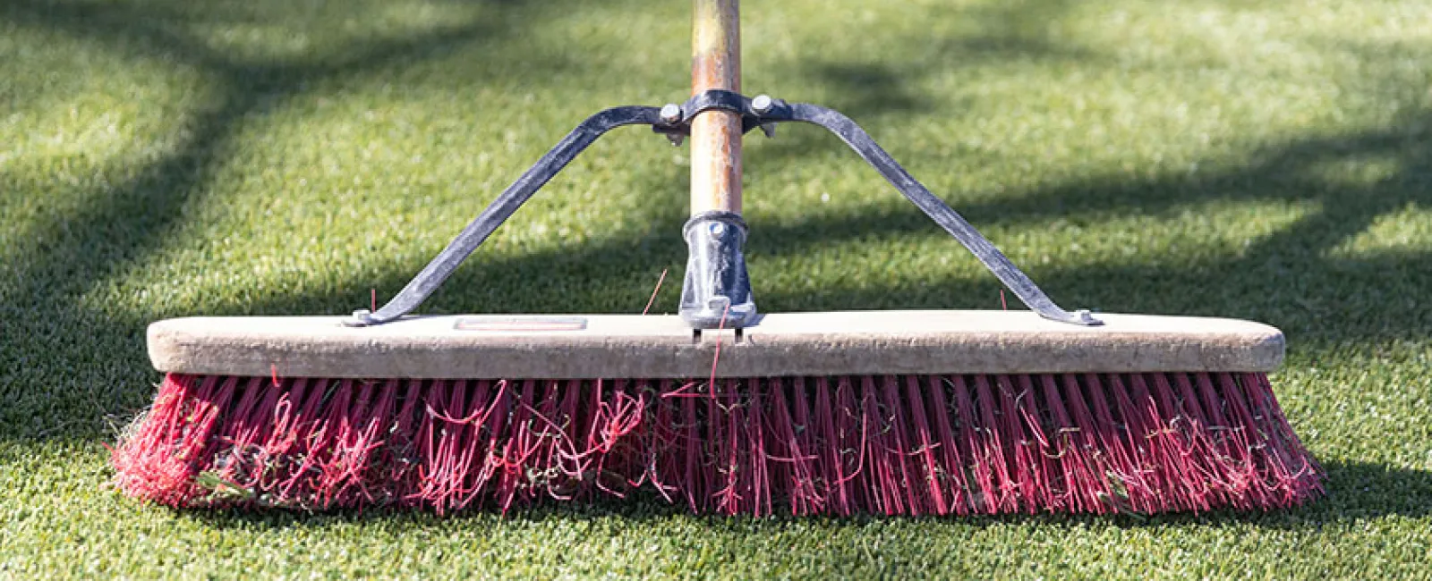 Close-up of a push broom with red bristles sweeping green artificial grass outdoors on a sunny day.