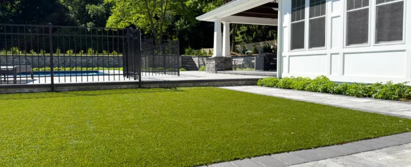 Modern backyard with artificial turf lawn, stone patio, white house, and black metal pool fence on a sunny day