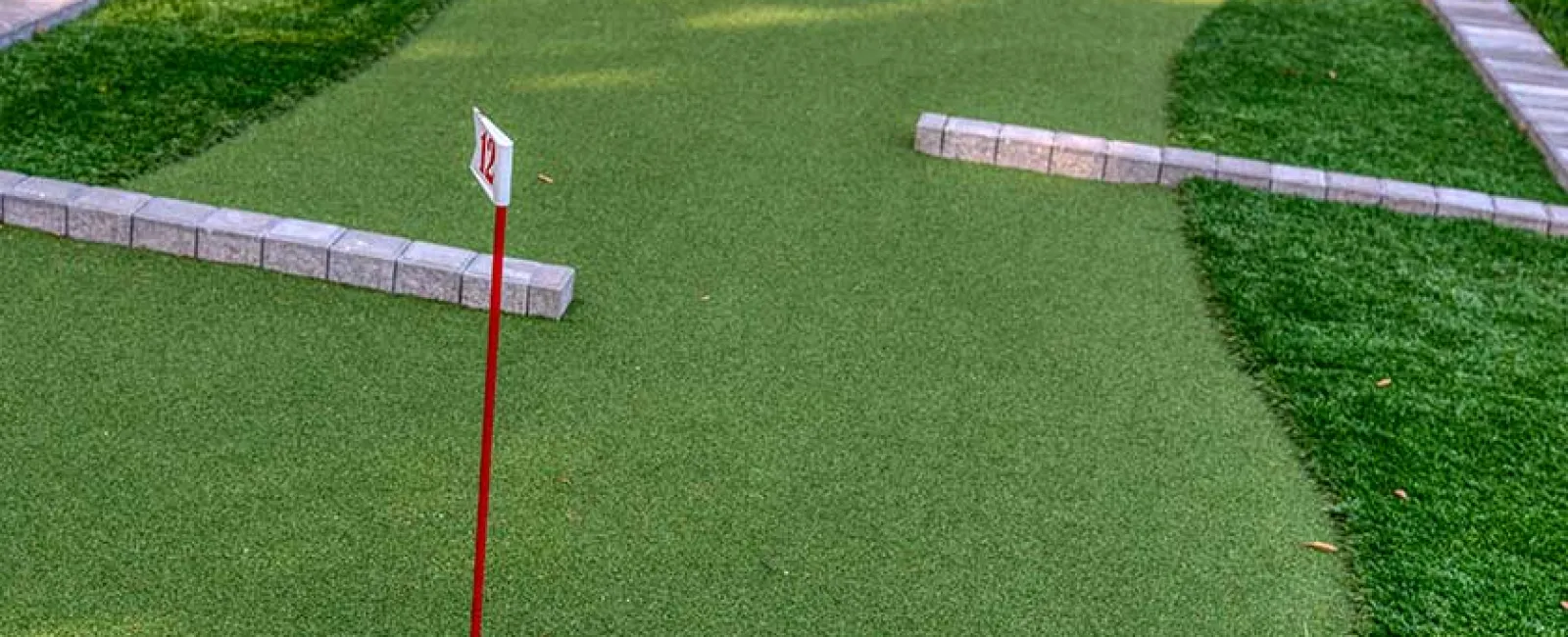 Miniature golf hole 12 with red flagpole and brick course boundaries on green artificial turf.