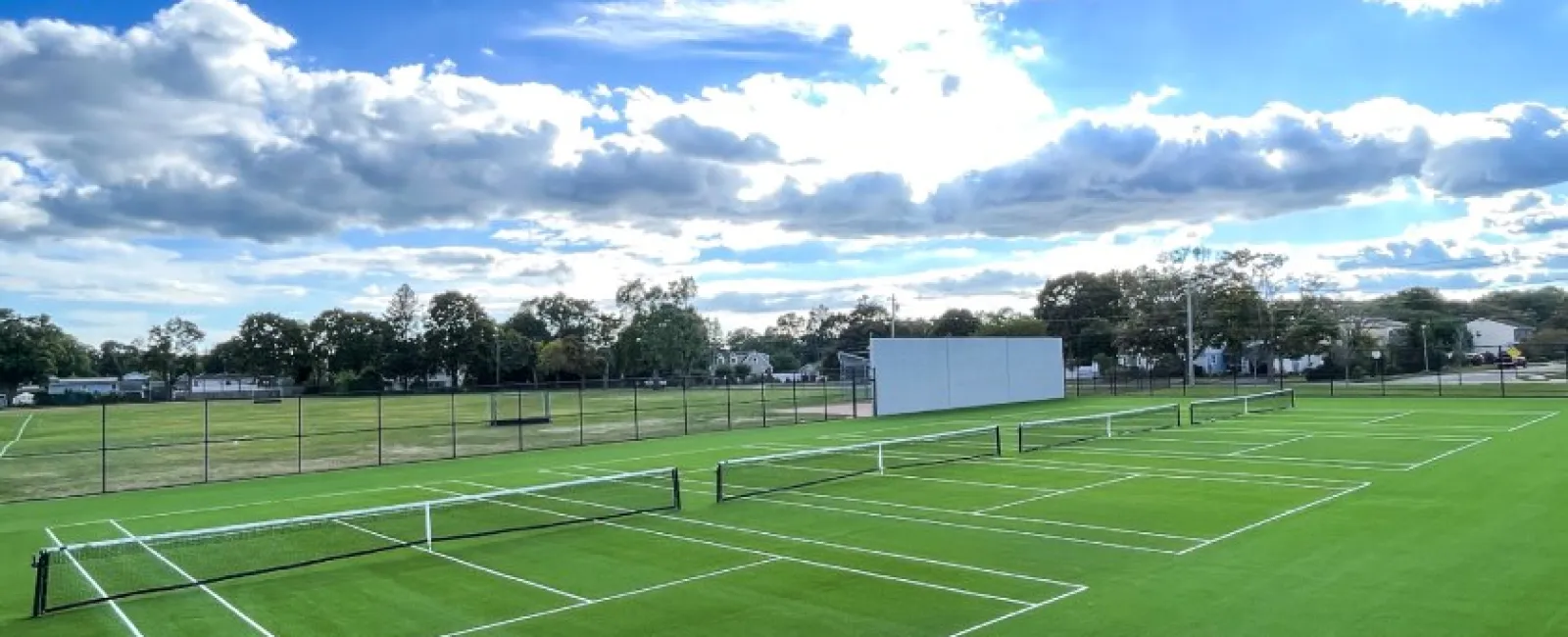 Empty green tennis courts under a bright sky with scattered clouds and surrounding trees and fences.