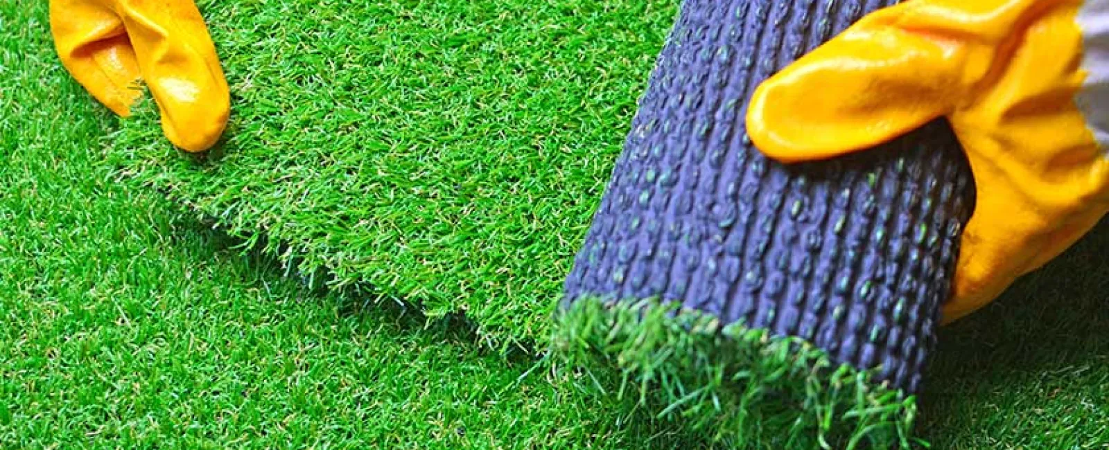 Hands in yellow gloves rolling out green artificial grass turf on a flat surface for installation.