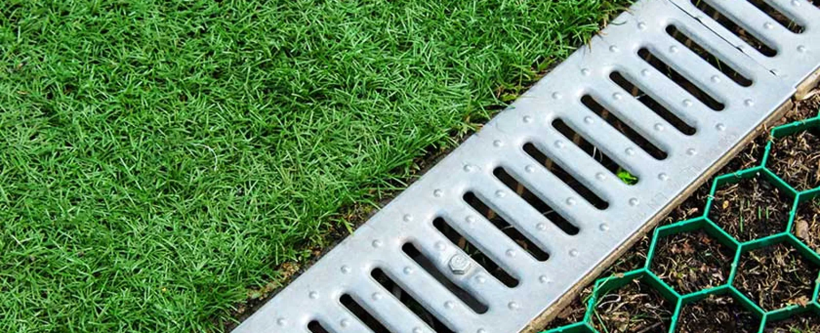 Green grass beside a metal drainage grate and hexagonal green mesh on soil.