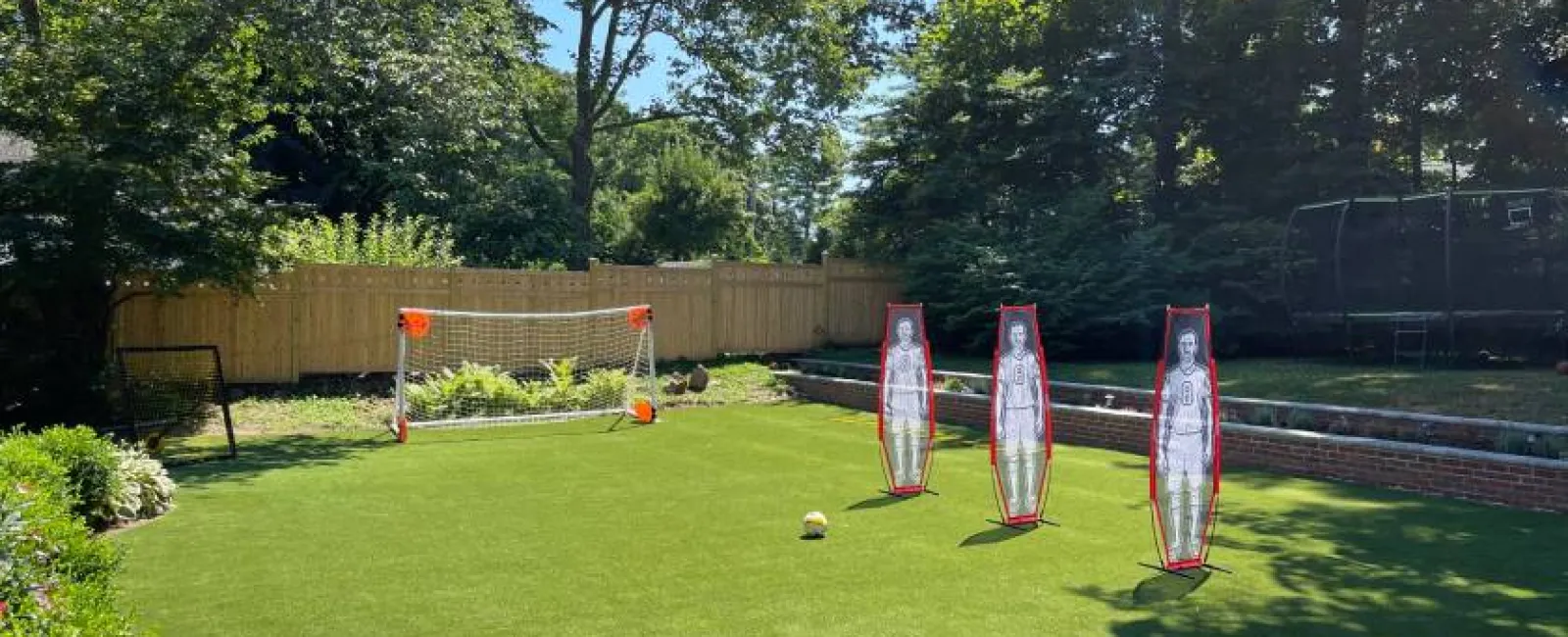 Backyard soccer practice setup with goal, three mannequin defenders, and soccer ball on green grass.