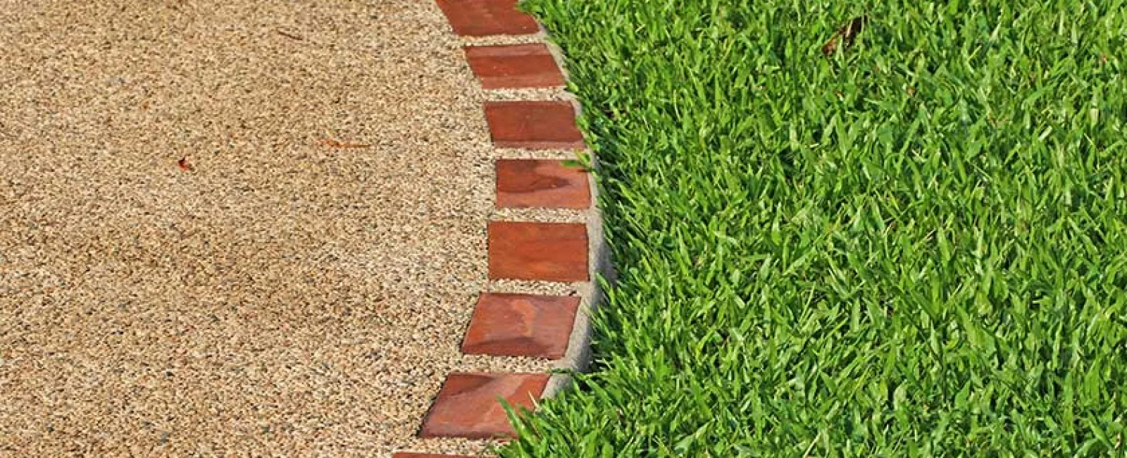 Curved edge of gravel pathway bordered by red bricks next to freshly cut green grass lawn