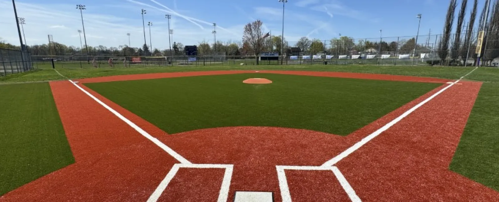 View of a baseball field from home plate showing red dirt, green grass, and clear blue sky.