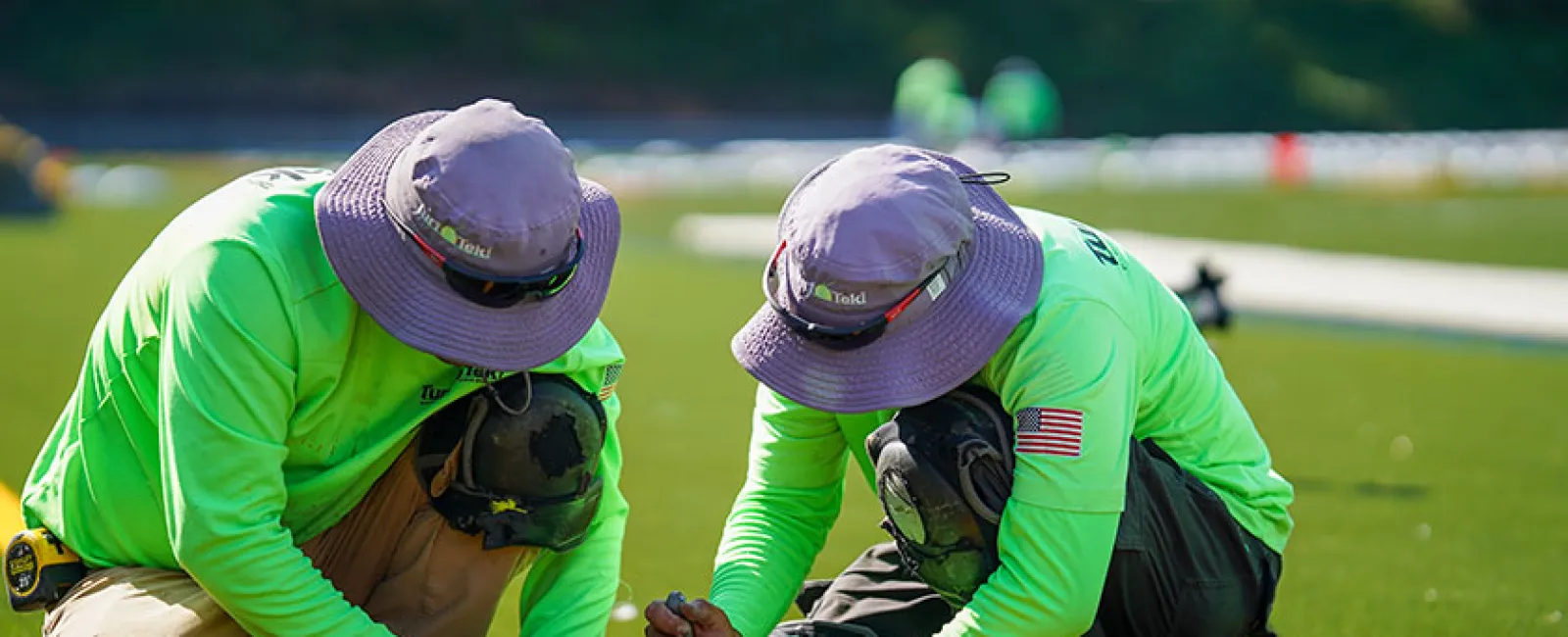 Two workers in bright green shirts and purple hats installing artificial turf on a sunny day outdoors