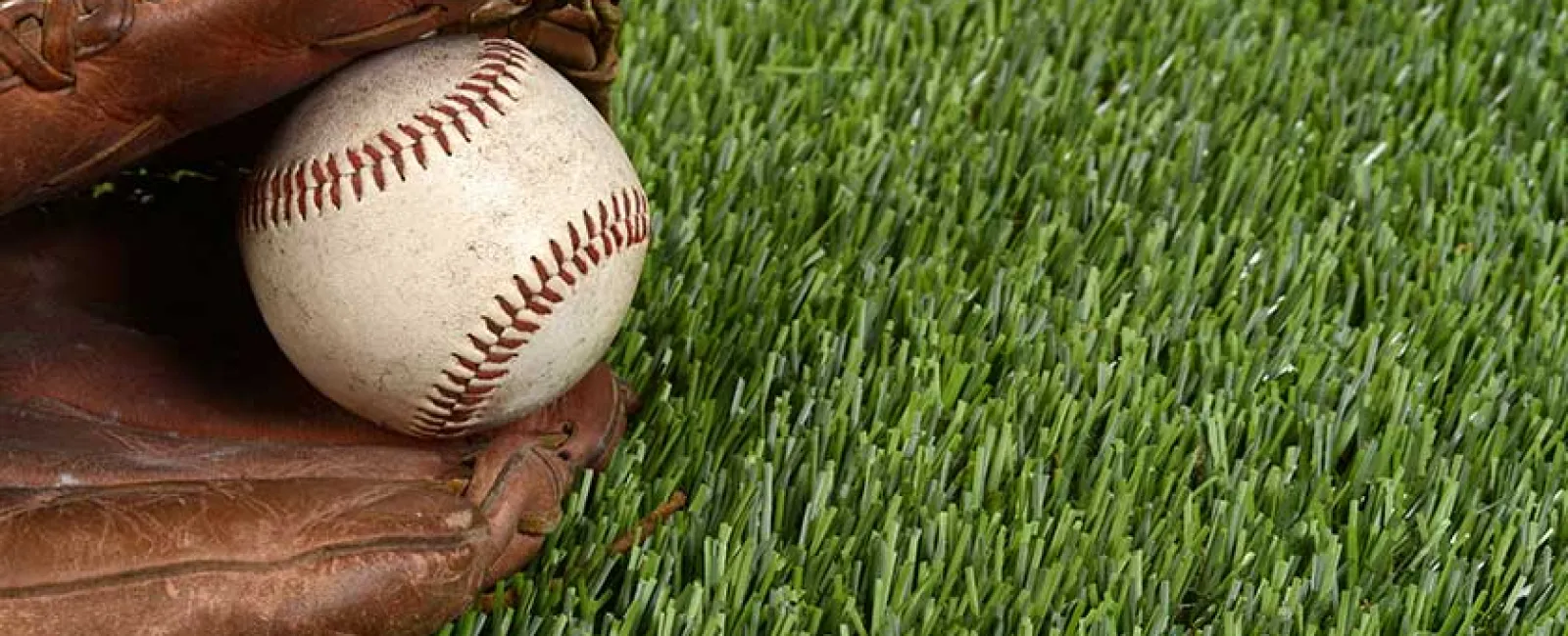 Baseball glove holding a worn baseball on green artificial grass field, close-up sports equipment image