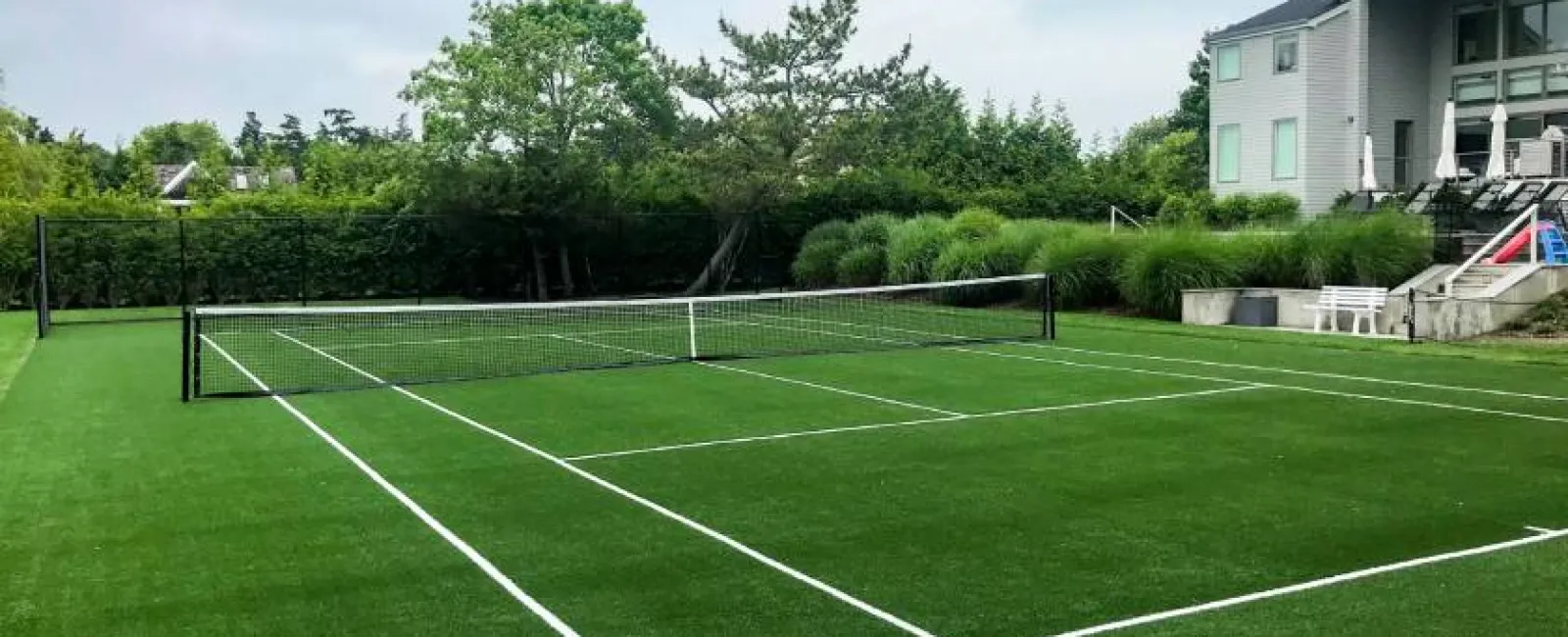 Residential backyard with a full-size artificial grass tennis court surrounded by greenery and a modern house.