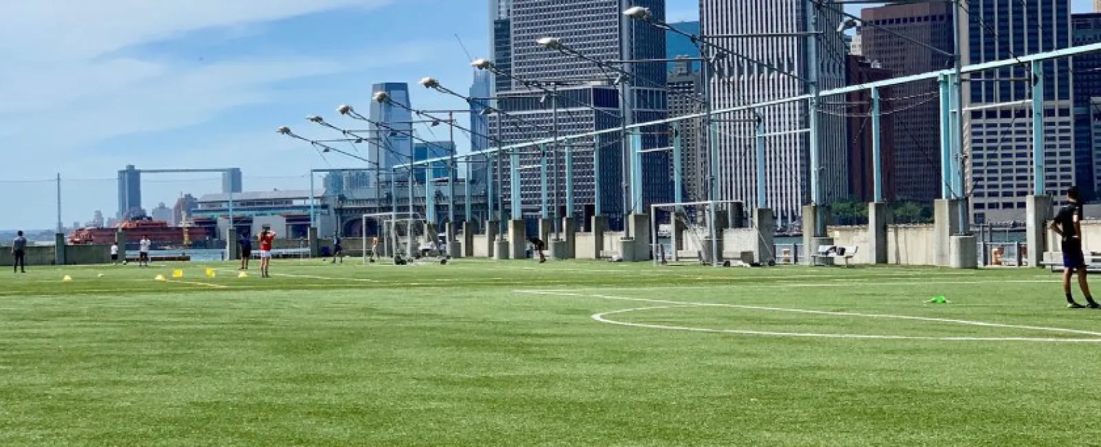Green soccer field with players and city skyline with tall buildings under blue sky in background