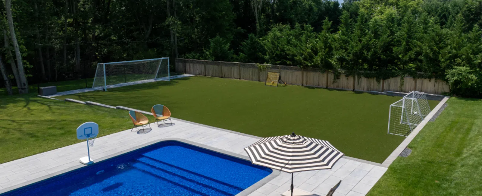 Backyard with blue pool, striped umbrellas, lounge chairs, basketball hoop, and soccer goals on artificial turf