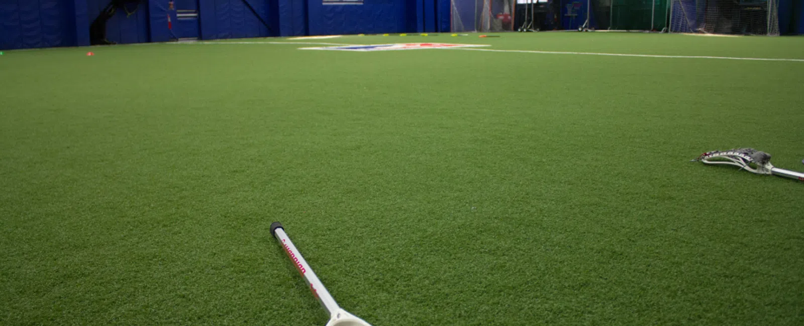 Indoor lacrosse field with green turf, two lacrosse sticks, and blue walls in the background