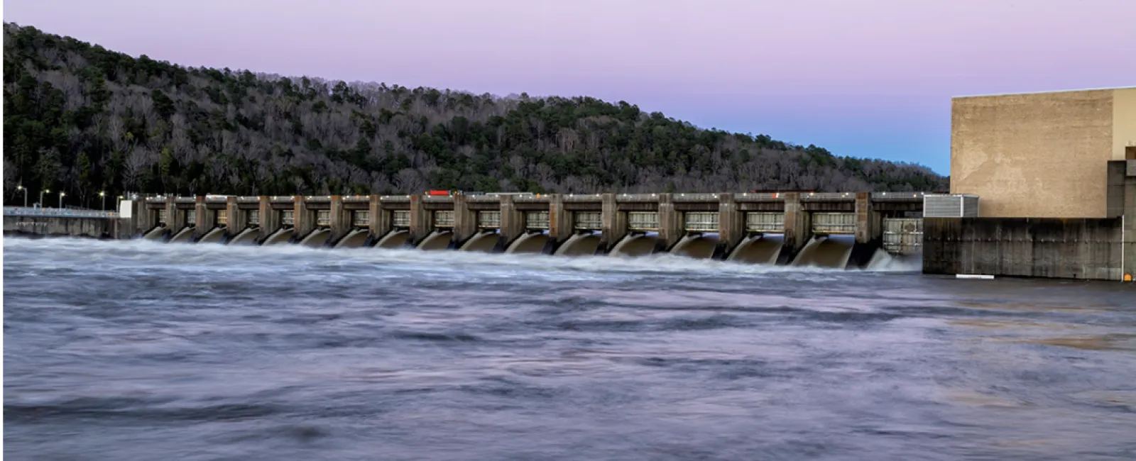 Hydroelectric dam releasing water with forested hill and clear sky in the background at dusk.