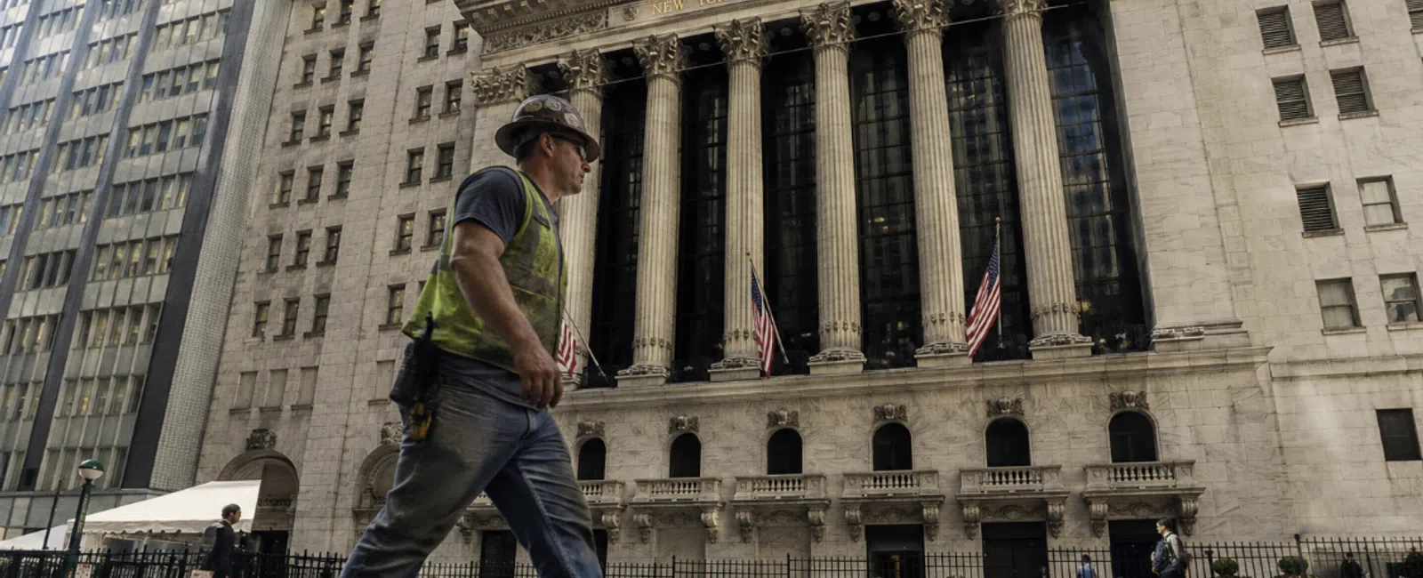 Construction worker walking past the iconic New York Stock Exchange building with columns and American flags.