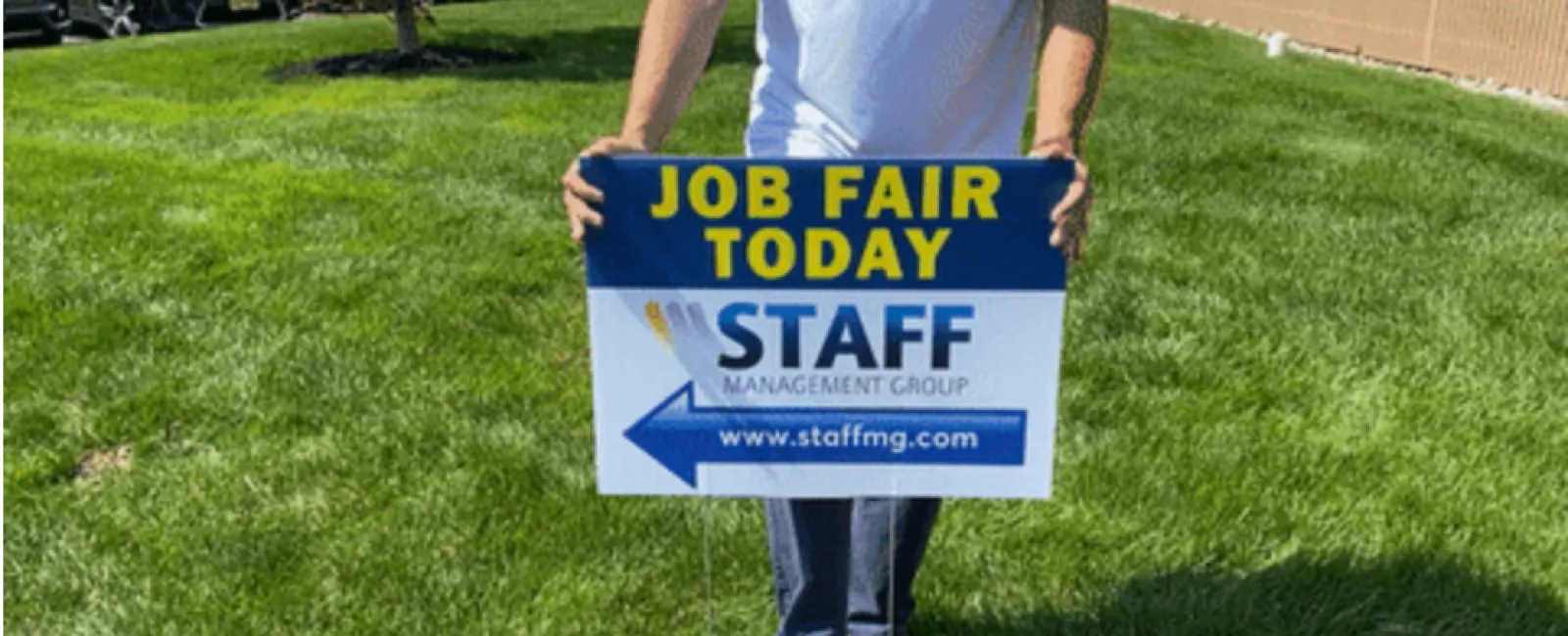 Person holding a Job Fair Today sign for Staff Management Group on green grass with blue sky.