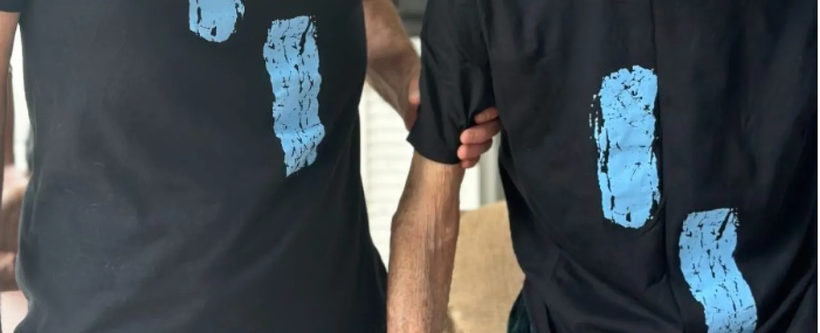 Two men wearing matching black t-shirts with blue footprints, smiling indoors in a room with bookshelves.
