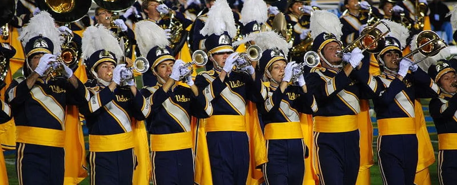 UCLA marching band members in navy and gold uniforms playing brass instruments during a stadium performance.