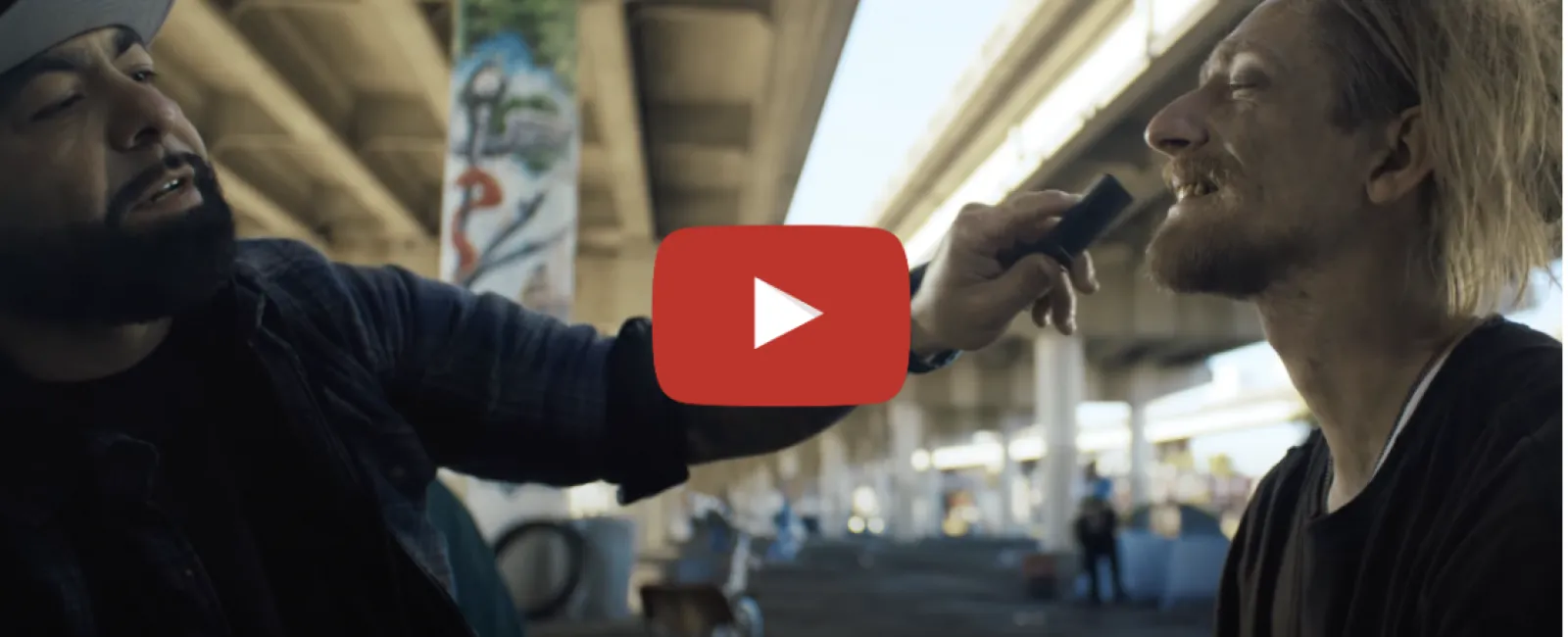 Man trimming another man’s beard under a graffiti-covered overpass in an urban setting.