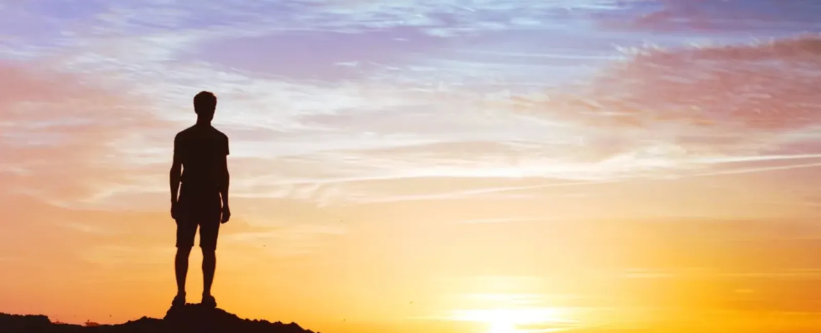 Silhouette of a person standing on rocky terrain watching a vibrant sunset over the ocean with colorful sky.