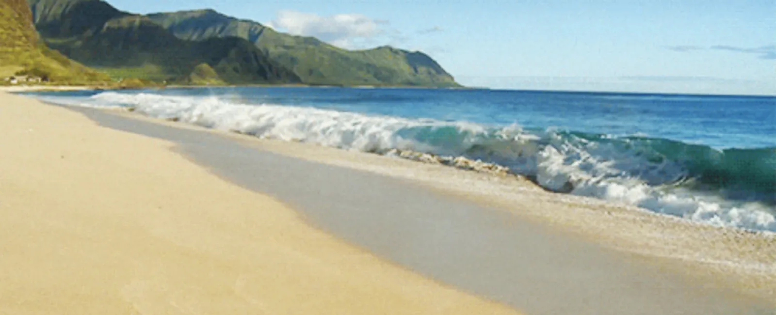 Sunny beach with gentle waves, golden sand, and green mountains under a clear blue sky.