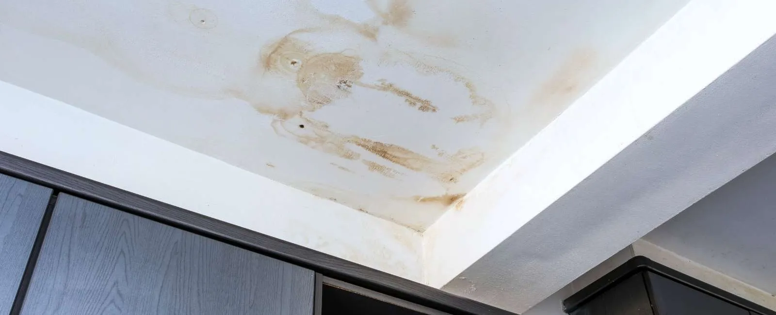Brown water stains and damage on a white ceiling corner above dark wooden cabinets indicating potential leak.