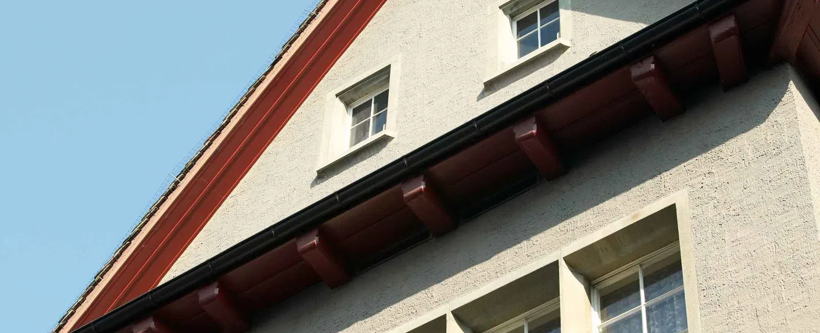 Close-up of a beige house gable with multiple windows and dark wooden eaves under a clear blue sky.