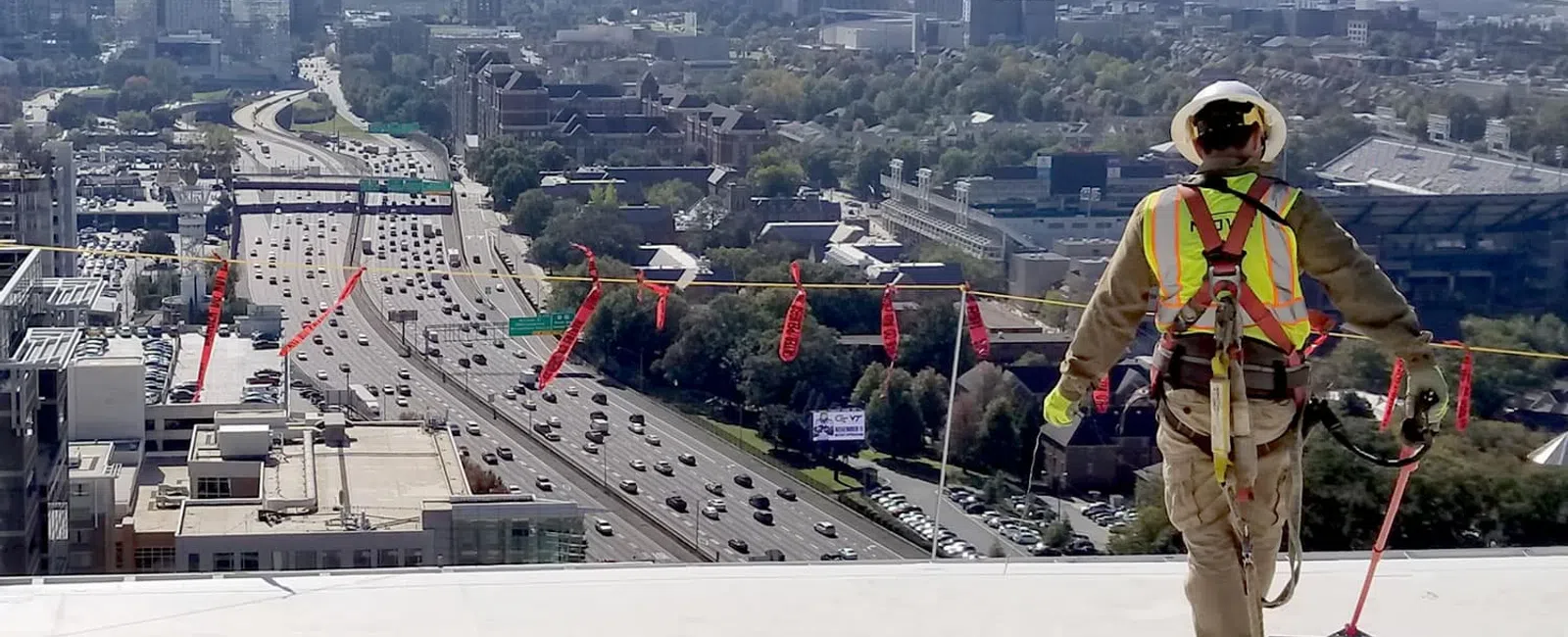 Construction worker in safety gear overlooking city skyline and busy highway from a high rooftop on a sunny day
