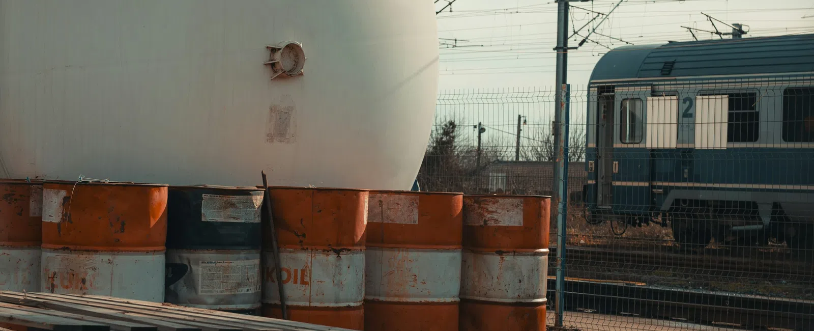 Large industrial storage tank with orange and white barrels, pipelines, and a train in the background on a cloudy day
