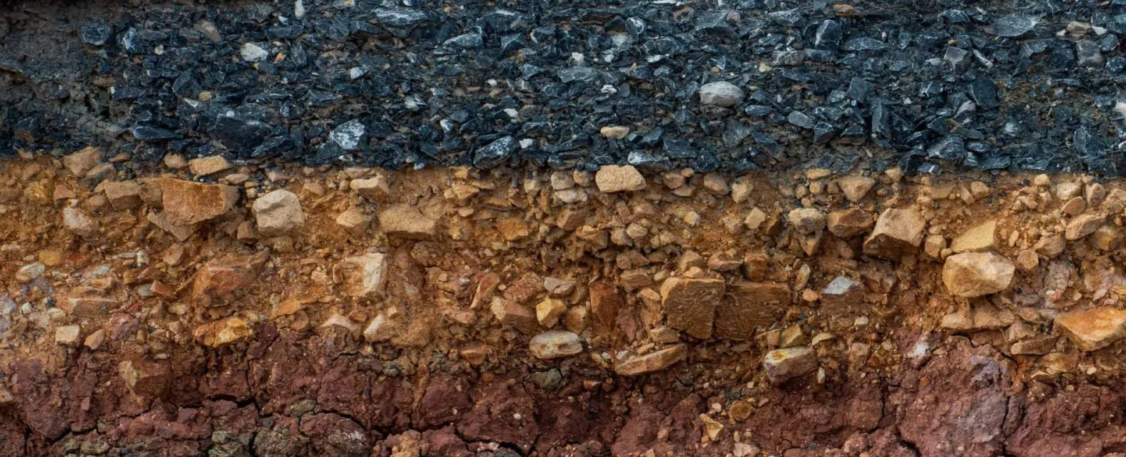 Cross-section of terrain showing multiple soil and rock layers topped by asphalt under a partly cloudy sky.