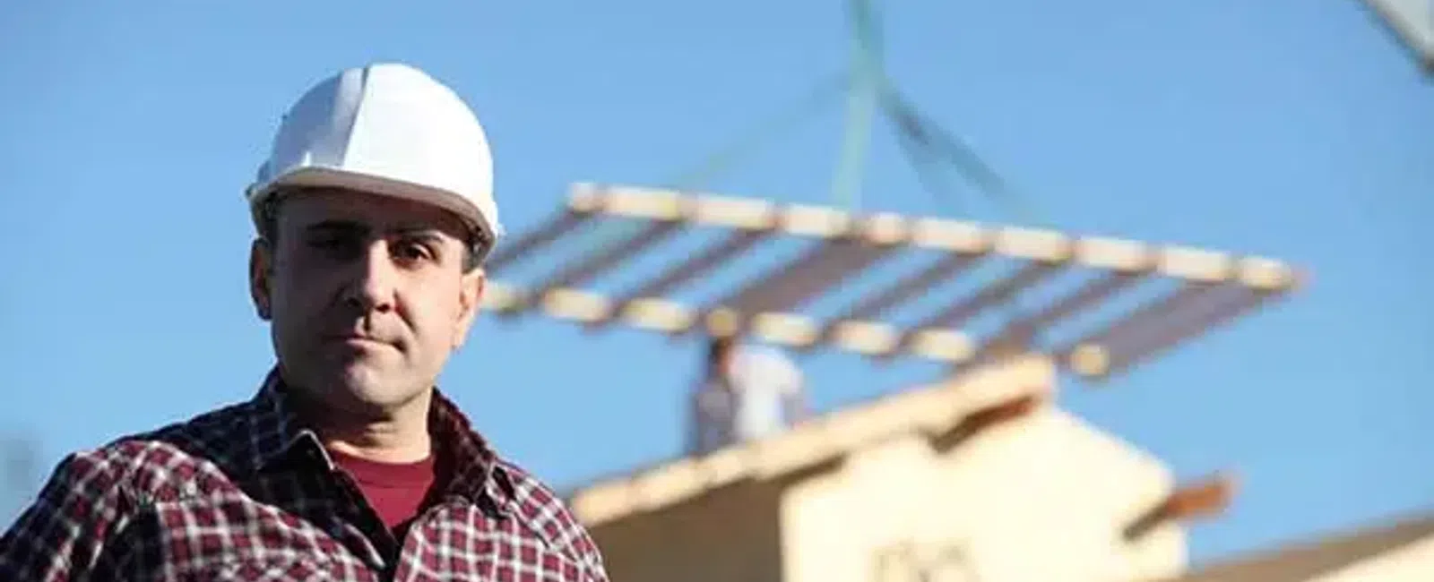 Construction worker wearing a white helmet and plaid shirt with building materials lifted by crane in background