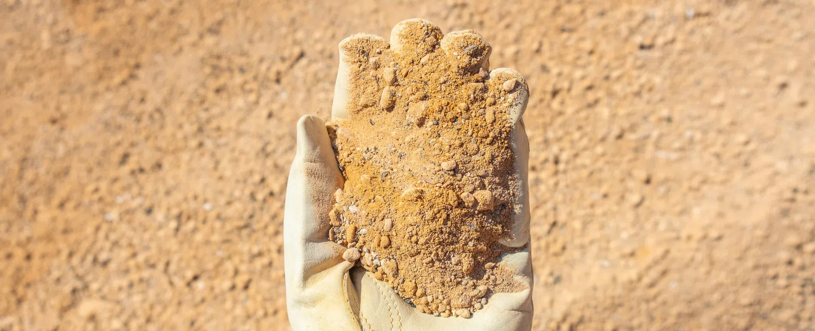 Gloved hand holding a handful of loose, sandy soil in front of a soil stockpile