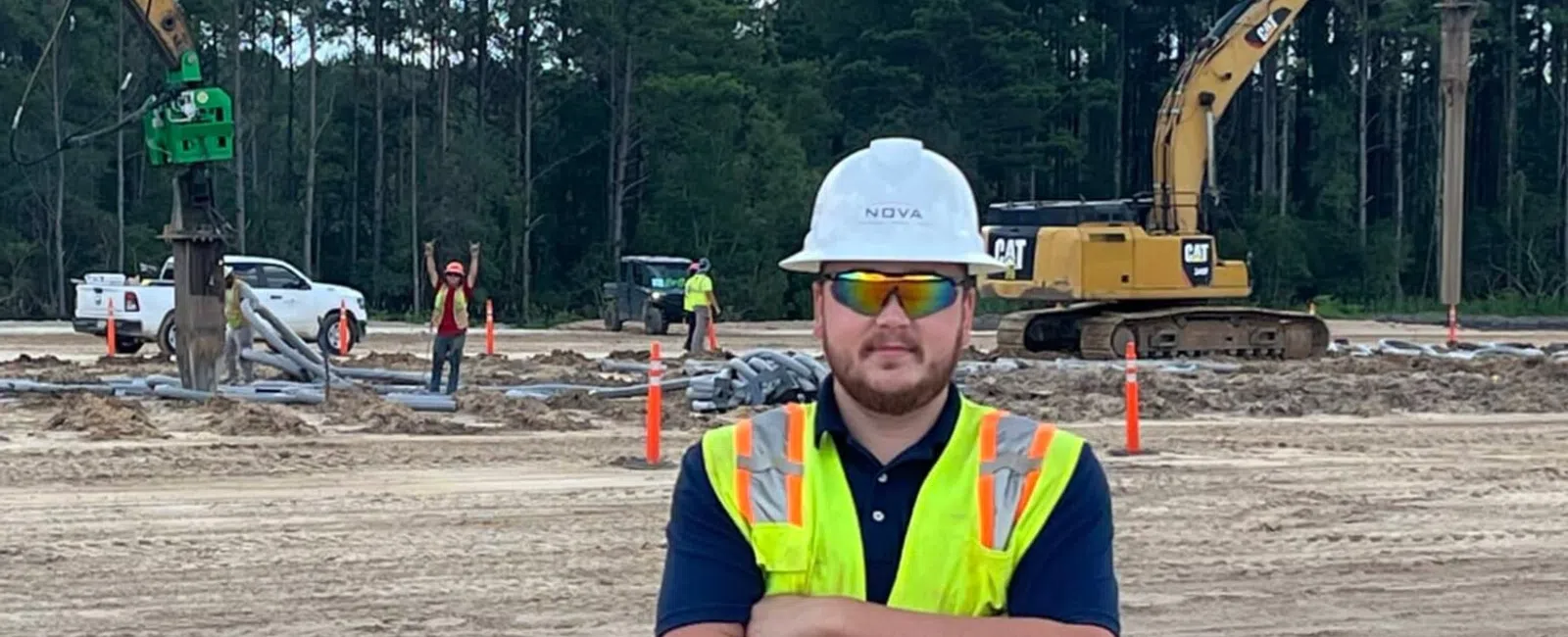 Construction worker in safety vest and helmet stands on site with excavators and forest background under blue sky.
