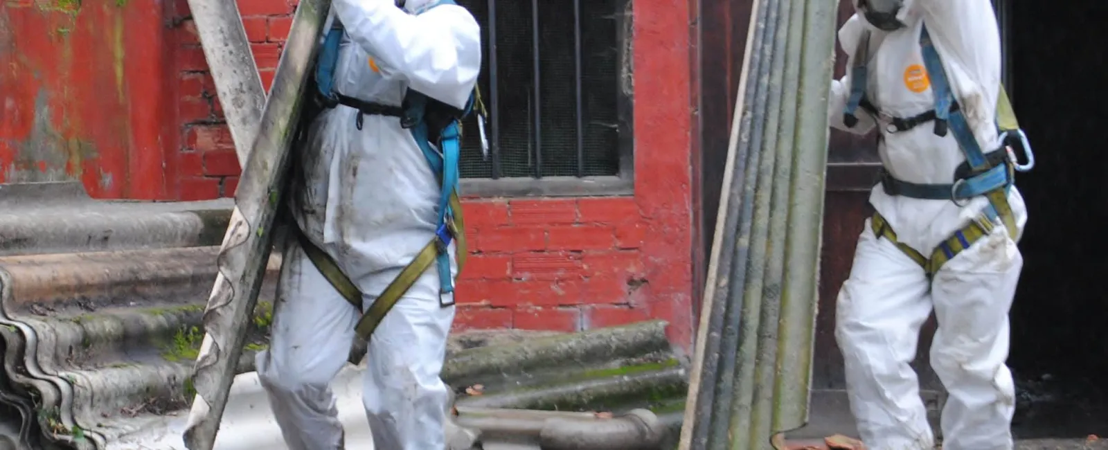 Two workers in protective suits and helmets carry corrugated asbestos sheets at a demolition site with a brick wall.