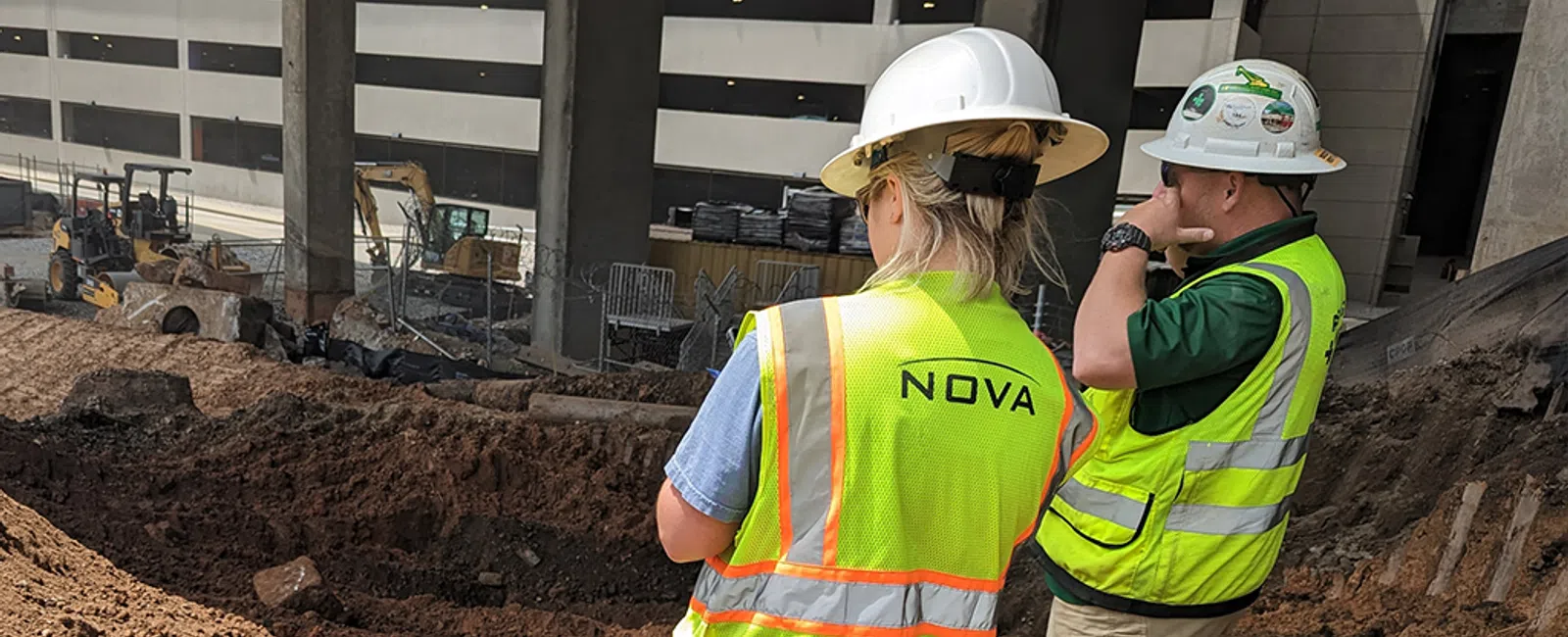 Two construction workers wearing hard hats and yellow vests standing on a dirt excavation site under a building.