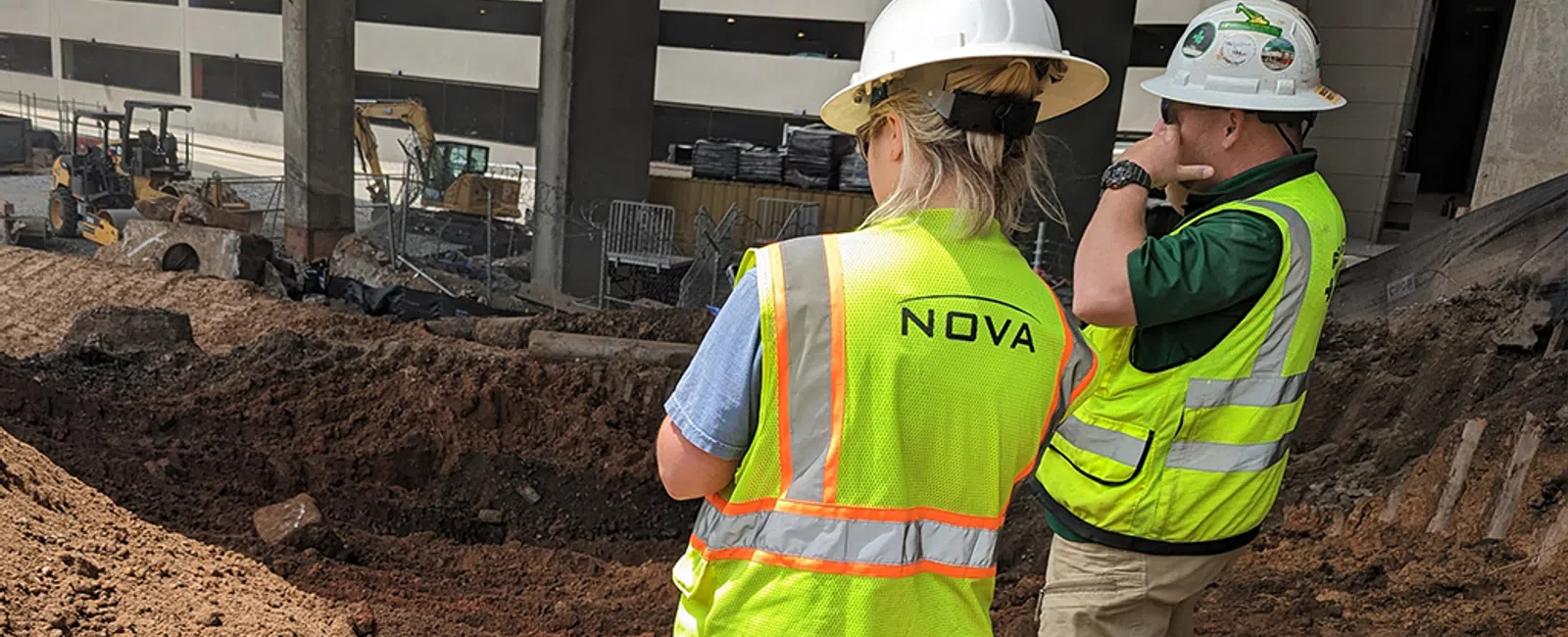Two construction workers wearing hard hats and yellow vests standing on a dirt excavation site under a building.