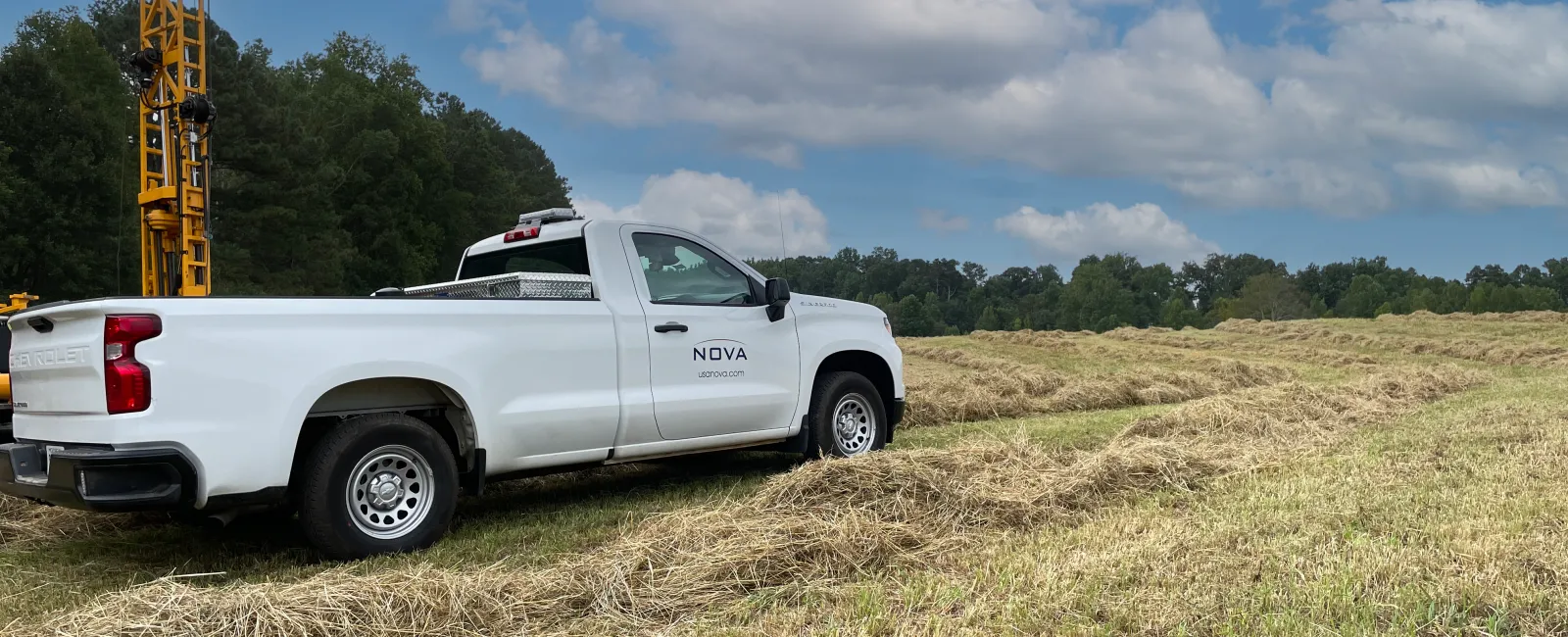 White NOVA pickup truck parked on a hay field with a drilling rig and trees under a partly cloudy sky.