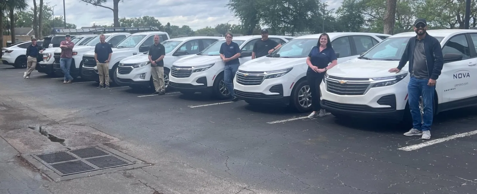 Team of eight people standing beside a row of eight identical white SUVs in a parking lot under cloudy sky.